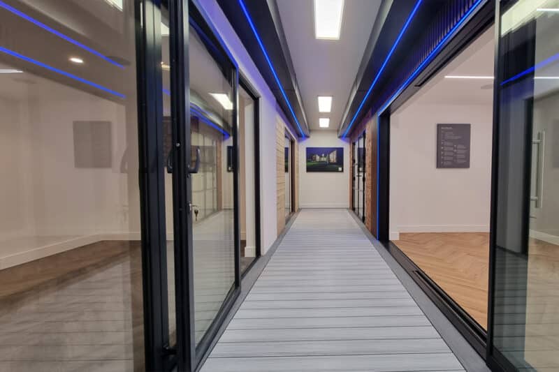 A modern corridor in Manchester features glass walls, blue LED strip lights on the ceiling, wooden flooring, and sleek sliding doors.