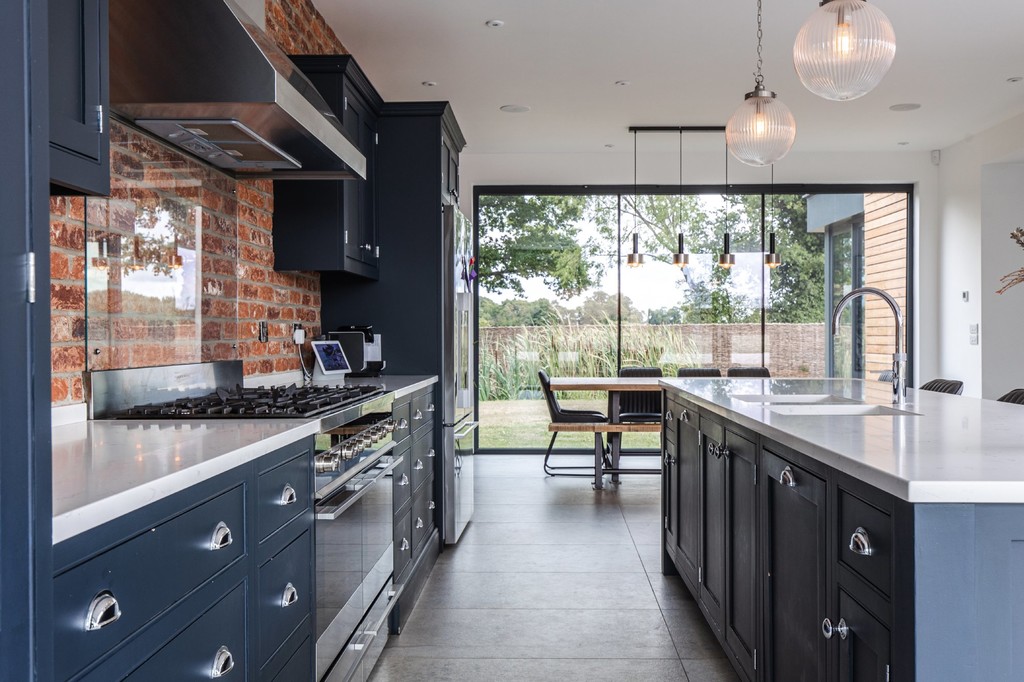 Modern kitchen with dark cabinets, large island, and stainless steel appliances. A brick wall accent, pendant lights, and a view of a dining table and outdoor greenery through glass doors.