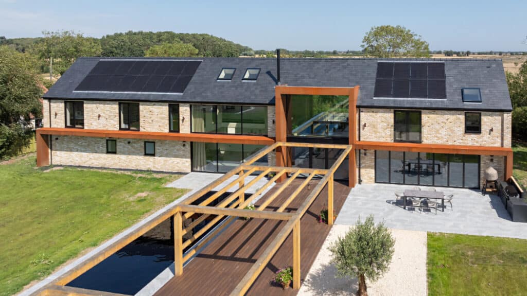 Modern two-story house with solar panels, stone and glass exterior, surrounded by green lawn. Features a wooden pergola and patio area with outdoor furniture.