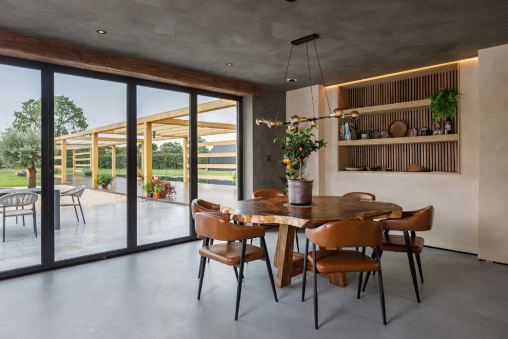 Modern dining room with a wooden table, six brown chairs, and a potted plant. Large glass doors open to a patio with outdoor seating and a view of greenery. Wood shelving with decor on the wall.