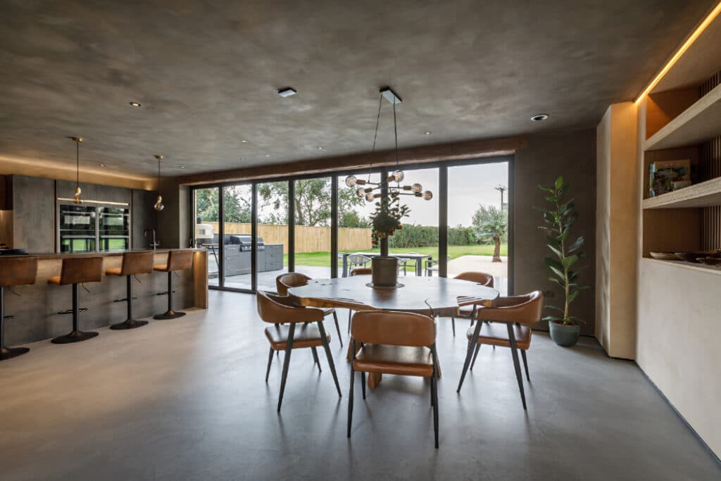 Modern kitchen with a round dining table, leather chairs, and a potted plant centerpiece. Large windows offer a view of the garden. Barstools line the kitchen counter on the left.