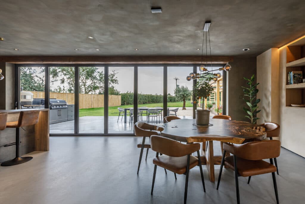 Modern dining area with a wooden table and chairs, large windows, and a view of a grassy yard with outdoor seating.