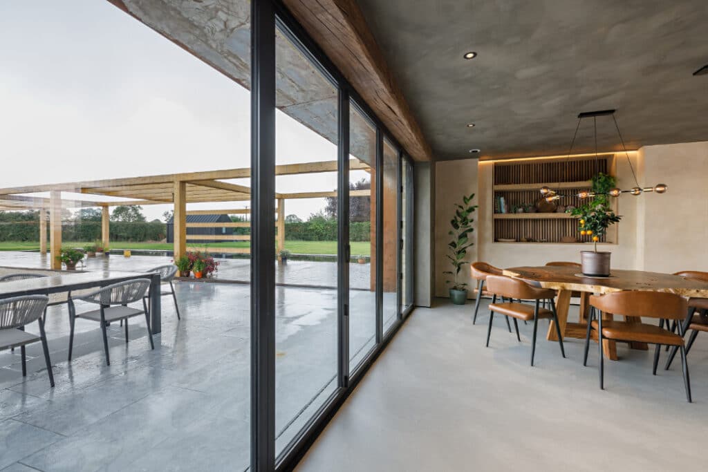 Modern dining area with wooden accents next to large glass doors opening to a patio with chairs and pergola. Greenery is visible outside.