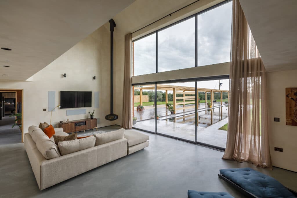 Spacious living room with large glass windows overlooking a patio and garden. Beige sectional sofa, mounted TV, and light curtains enhance a minimalist design with high ceilings.