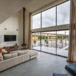 Thumbnail of http://Spacious%20living%20room%20with%20large%20glass%20windows%20overlooking%20a%20patio%20and%20garden.%20Beige%20sectional%20sofa,%20mounted%20TV,%20and%20light%20curtains%20enhance%20a%20minimalist%20design%20with%20high%20ceilings.