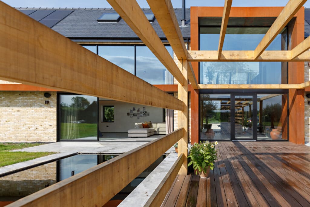 Modern house with large glass windows and wooden beams overhead. A patio with wooden flooring and a potted plant leads to the entrance.