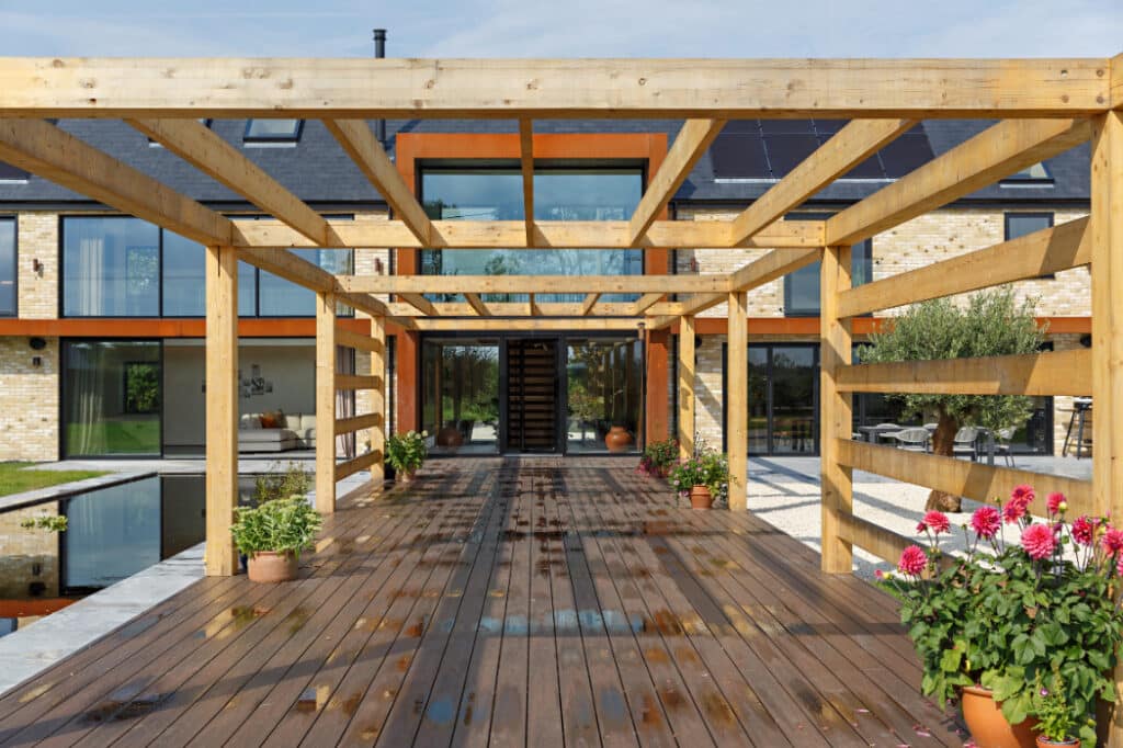 Wooden pergola over a wet deck leading to a modern two-story brick house with large windows. Potted plants line the walkway.