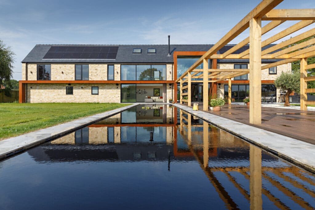 Modern house with solar panels and large windows, featuring a reflective rectangular pool and a wooden pergola in the foreground, set in a grassy yard under a clear blue sky.