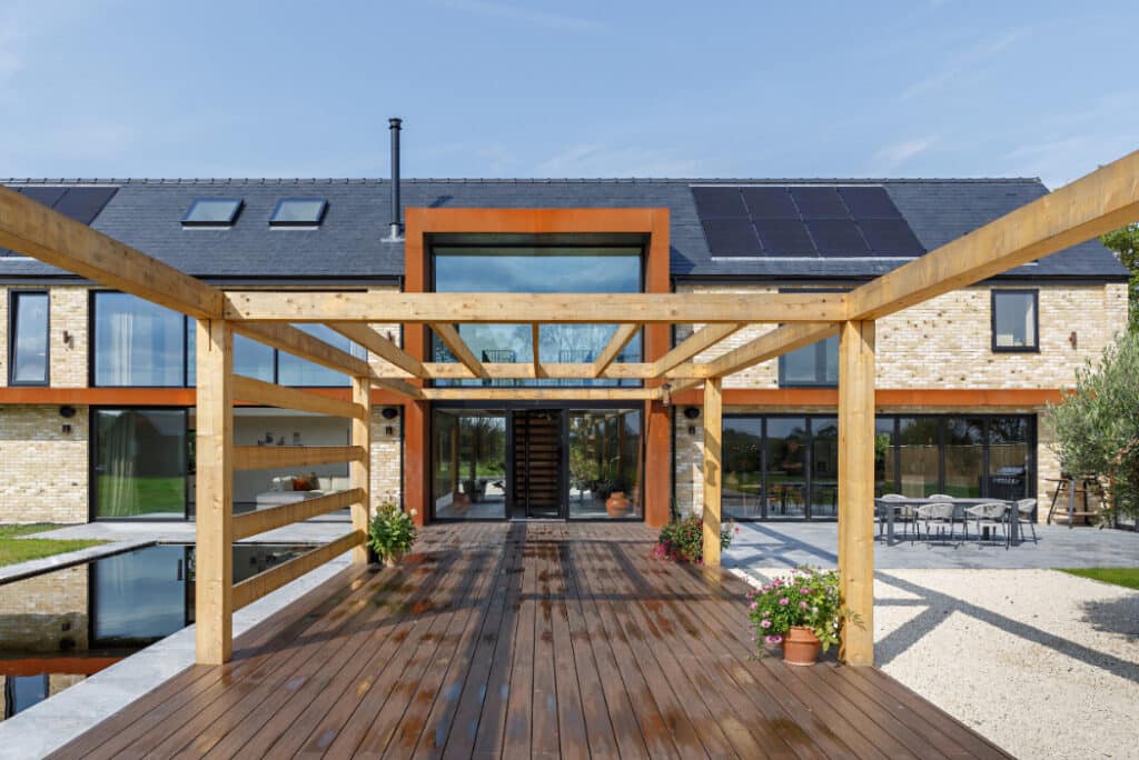 Modern two-story house with solar panels, a wooden pergola on a deck, and glass doors. Reflective water feature and patio chairs are visible in the foreground.