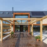 Thumbnail of http://Modern%20two-story%20house%20with%20solar%20panels,%20a%20wooden%20pergola%20on%20a%20deck,%20and%20glass%20doors.%20Reflective%20water%20feature%20and%20patio%20chairs%20are%20visible%20in%20the%20foreground.