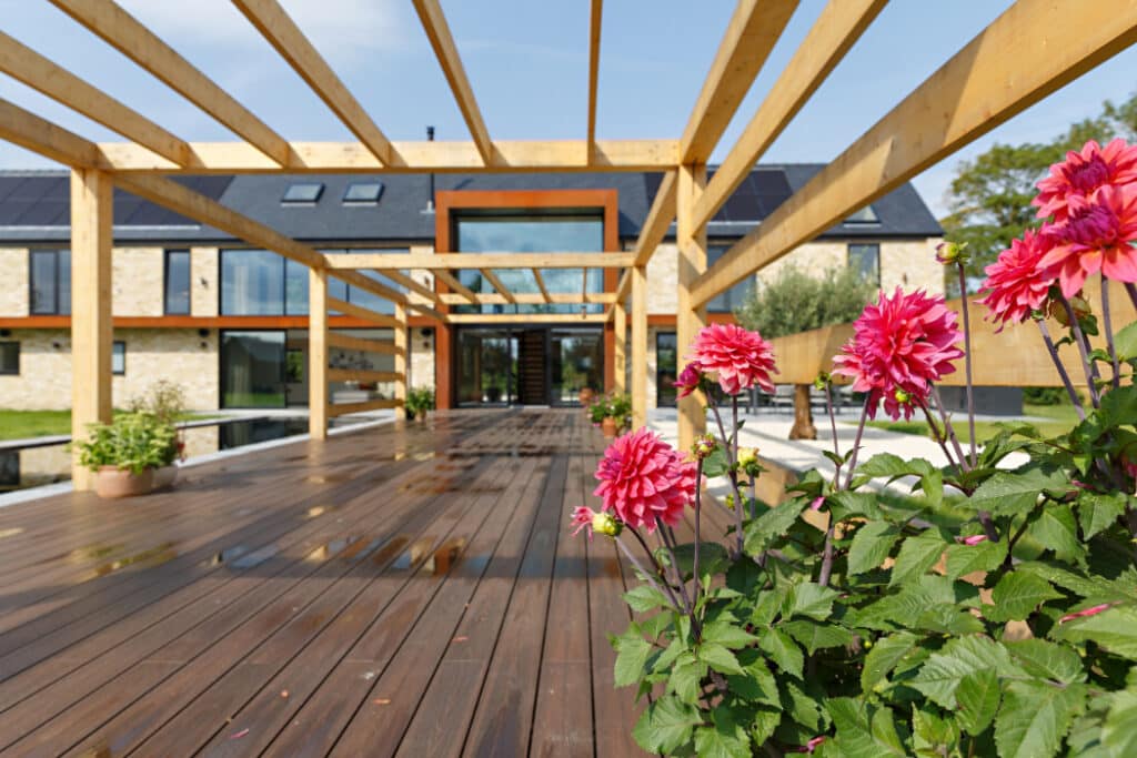Contemporary house with exposed wooden beams, large glass doors, and a wooden deck. Pink flowers are in the foreground.