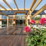Thumbnail of http://Contemporary%20house%20with%20exposed%20wooden%20beams,%20large%20glass%20doors,%20and%20a%20wooden%20deck.%20Pink%20flowers%20are%20in%20the%20foreground.