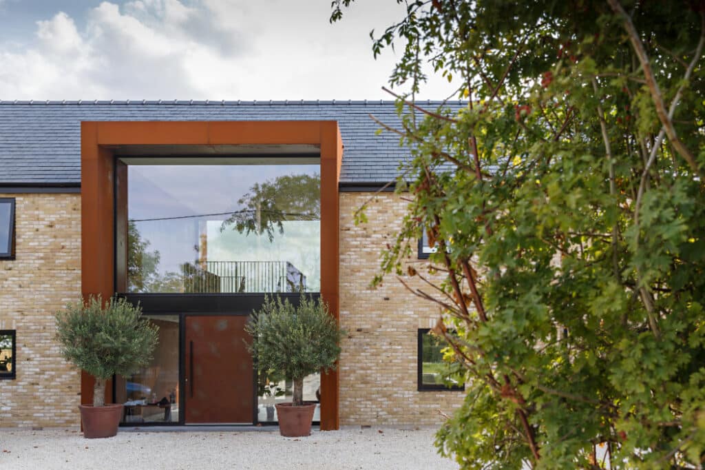 Modern brick house with large glass entrance and rust-colored frame, flanked by two potted trees. Sparse gravel yard in foreground and leafy tree branch on the right.