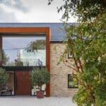 Thumbnail of http://Modern%20brick%20house%20with%20large%20glass%20entrance%20and%20rust-colored%20frame,%20flanked%20by%20two%20potted%20trees.%20Sparse%20gravel%20yard%20in%20foreground%20and%20leafy%20tree%20branch%20on%20the%20right.