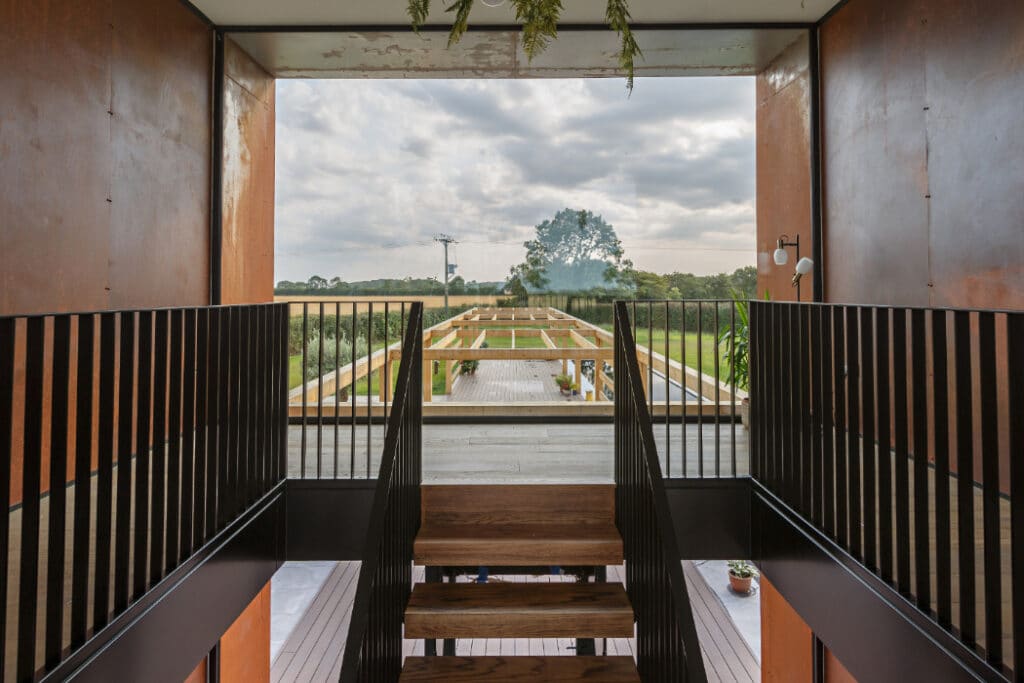 Staircase in a modern building leads to an outdoor terrace with a view of a grassy landscape, trees, and a wind turbine in the distance.