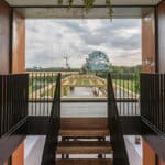 Thumbnail of http://Staircase%20in%20a%20modern%20building%20leads%20to%20an%20outdoor%20terrace%20with%20a%20view%20of%20a%20grassy%20landscape,%20trees,%20and%20a%20wind%20turbine%20in%20the%20distance.