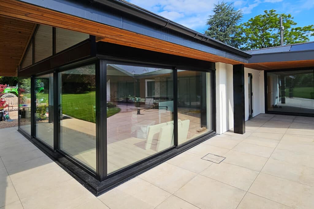 Modern house exterior with large glass windows, black frames, and a beige tiled patio. Visible interior features white sofas. Green lawn and trees in the background under a blue sky.