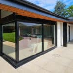 Thumbnail of http://Modern%20house%20exterior%20with%20large%20glass%20windows,%20black%20frames,%20and%20a%20beige%20tiled%20patio.%20Visible%20interior%20features%20white%20sofas.%20Green%20lawn%20and%20trees%20in%20the%20background%20under%20a%20blue%20sky.