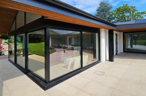 Modern house exterior with large glass windows, black frames, and a beige tiled patio. Visible interior features white sofas. Green lawn and trees in the background under a blue sky.