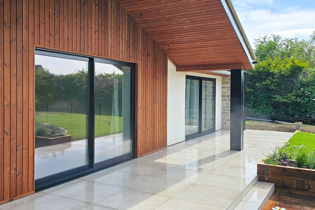 Modern house exterior with wooden siding and large glass sliding doors. A polished stone patio extends to a grassy lawn, bordered by a raised garden bed.