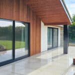 Thumbnail of http://Modern%20house%20exterior%20with%20wooden%20siding%20and%20large%20glass%20sliding%20doors.%20A%20polished%20stone%20patio%20extends%20to%20a%20grassy%20lawn,%20bordered%20by%20a%20raised%20garden%20bed.