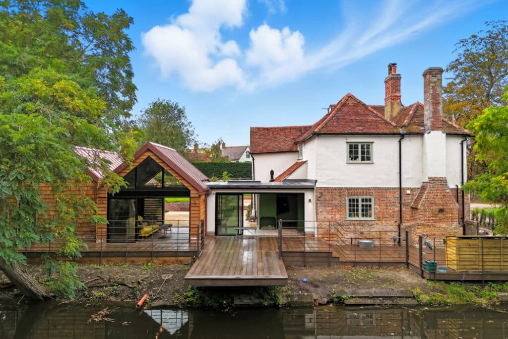 A riverside house with a mix of modern and traditional architecture, featuring large glass doors, wooden decking, brick walls, and a red-tiled roof, surrounded by trees.