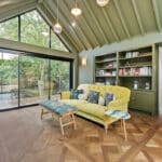 Thumbnail of http://Contemporary%20sunroom%20with%20Sunflex%20bifold%20doors%20opening%20onto%20terrace,%20yellow%20sofa,%20green%20built-in%20shelving,%20herringbone%20flooring,%20and%20vaulted%20ceiling%20with%20skylights.