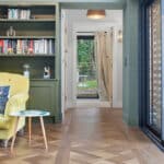 Thumbnail of http://Historic%20home%20interior%20corridor%20with%20black%20bifold%20doors%20opening%20to%20garden,%20yellow%20accent%20chair,%20bookshelves,%20and%20herringbone%20wood%20flooring.