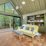 Thumbnail of http://Modern%20living%20room%20with%20yellow%20sofa,%20olive-green%20built-in%20shelving,%20and%20expansive%20glazed%20bifold%20doors%20opening%20to%20tree-lined%20deck%20overlooking%20countryside.