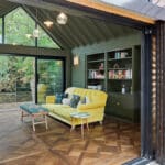 Thumbnail of http://Sunflex%20bifold%20doors%20open%20contemporary%20living%20space%20to%20woodland%20views,%20featuring%20yellow%20sofa,%20built-in%20shelving,%20herringbone%20flooring%20and%20gable%20roof%20lighting.