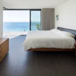 Thumbnail of http://Myrio%20Sennen%20Cove%20bedroom%20with%20expansive%20glazed%20bifold%20doors%20framing%20coastal%20sea%20views,%20featuring%20dark%20flooring,%20timber%20furnishings,%20and%20minimalist%20design.