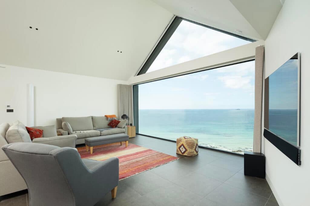 Modern living room with grey sofas, a red rug, and a large window offering a clear view of the sea and sky. A flat-screen TV is mounted on the right wall.